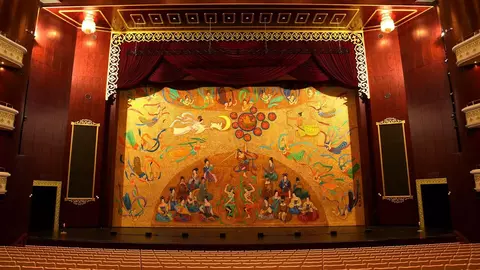 Theater stage featuring a colorful, abstract backdrop with dancers and musicians, framed by red curtains and ornate detailing.