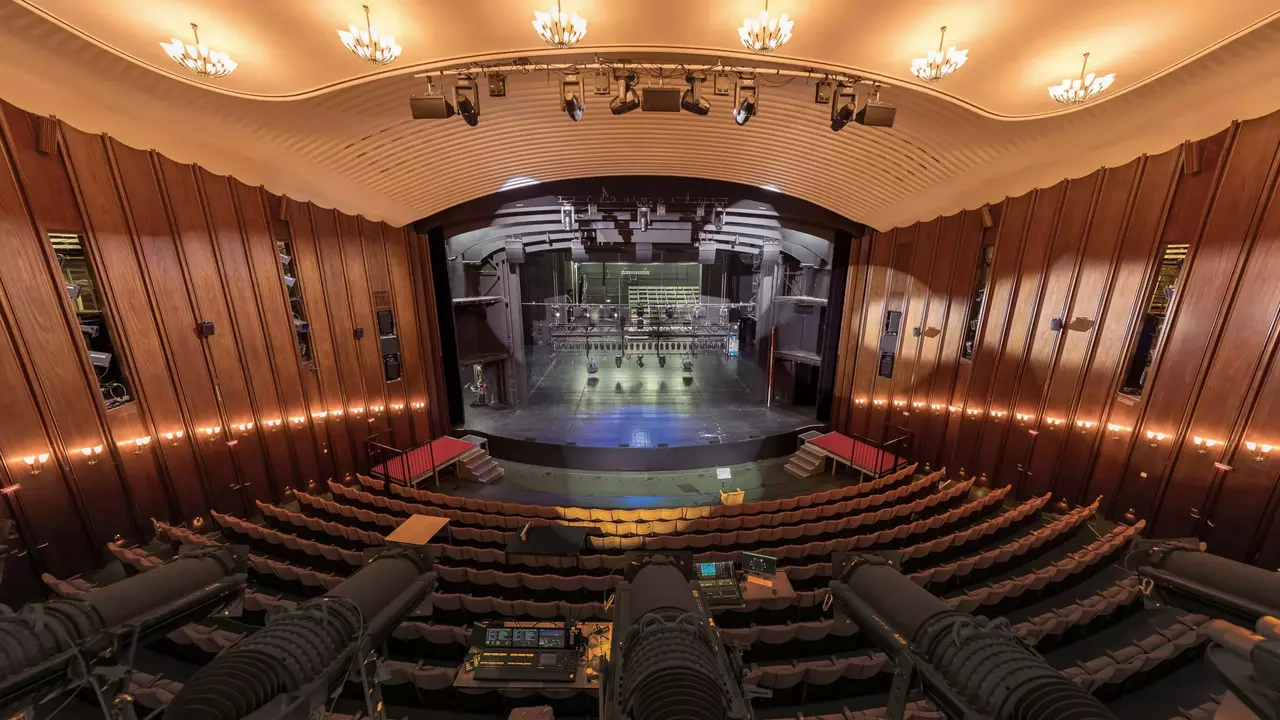 The interior of a theater featuring a stage with lighting equipment, rows of seats, and wooden paneling along the walls.