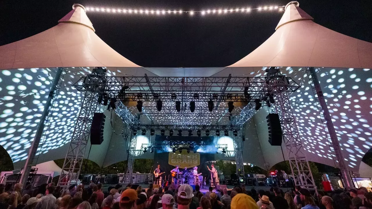 A band performs on stage under illuminated tents, with a crowd of spectators enjoying the concert at night.