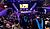 Graduates in caps and gowns walk down the stairs towards a stage decorated with lights and a large screen, while an audience watches.