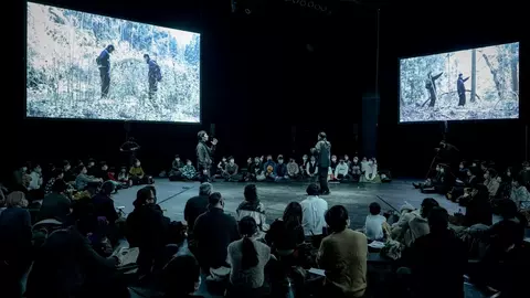 A performance taking place on stage with two large screens displaying scenes in the background, surrounded by an audience seated on the floor.