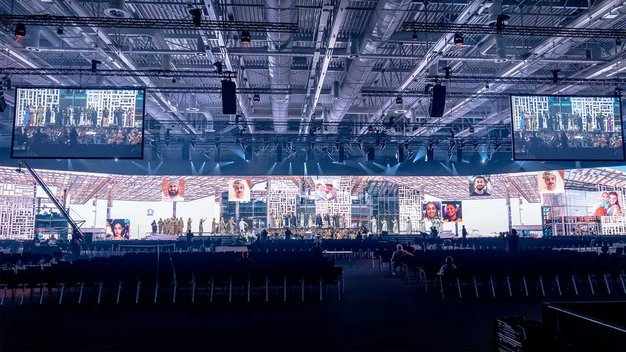 Large indoor event space with a stage, multiple screens displaying images, and rows of empty seats.