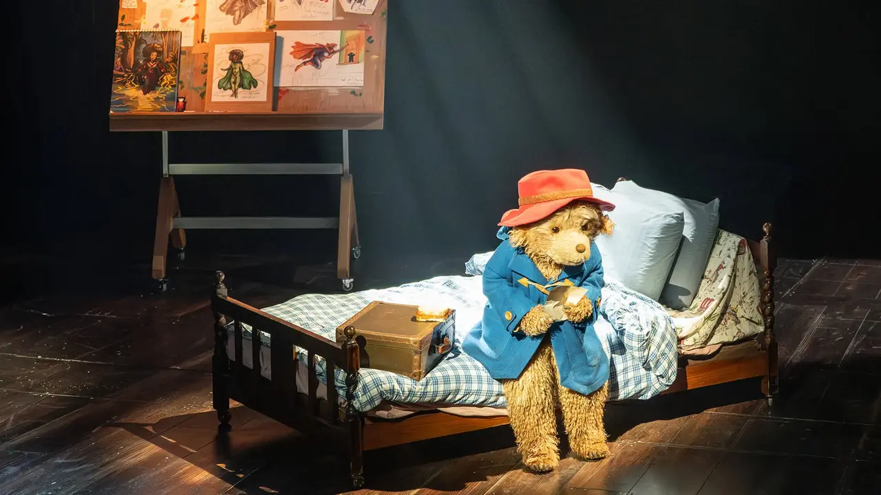 A teddy bear in a blue coat and red hat sits on a bed, illuminated by a spotlight, with a board of drawings in the background.