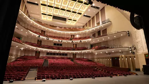 Interior view of a modern theater with multiple seating levels, featuring red seats and a well-lit ceiling.