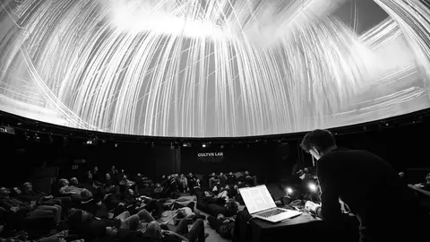 Audience lying on the floor of a dome theater, watching a light display projected on the ceiling, with a presenter at a table.
