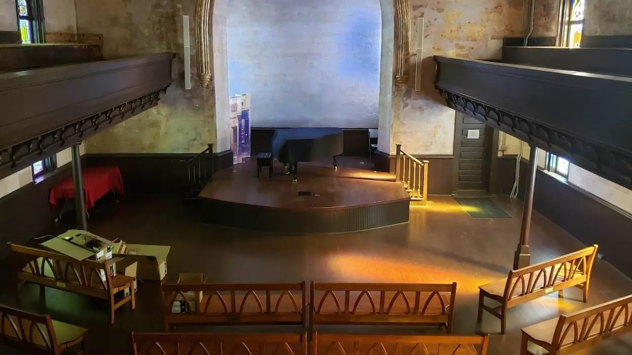 Interior of a historic hall featuring a raised stage, wooden seating, and stained glass windows, with a simple, rustic design.