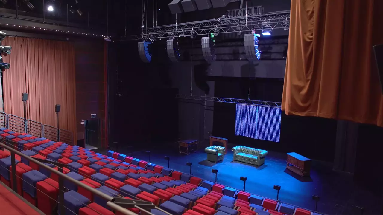 Leeres Theater mit roten und blauen Sitzplätzen, ausgestattet mit einer Bühne, auf der zwei grüne Sofas und ein Tisch stehen, beleuchtet von Bühnenlichtern.