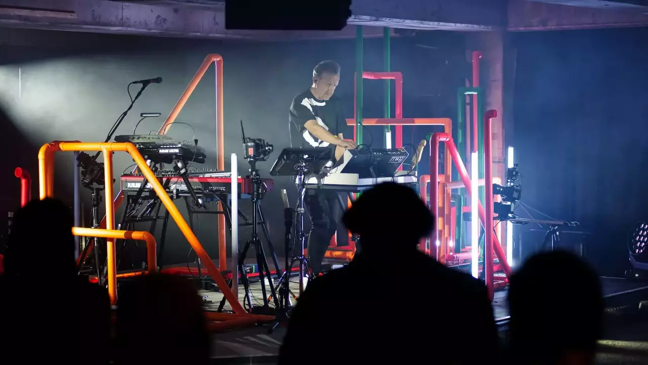 A musician performs on stage surrounded by colorful geometric structures and electronic instruments, with a dimly lit audience in the foreground.