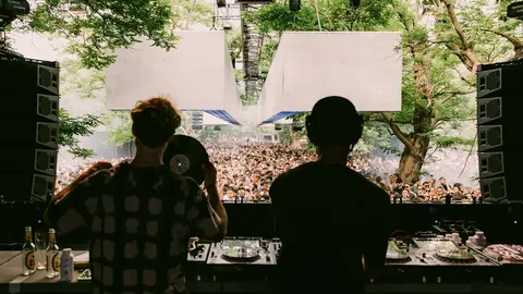 Two DJs perform at a music festival, facing a large crowd under trees, with sound equipment visible in the foreground.