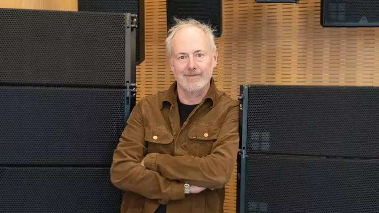 A man with gray hair stands confidently with arms crossed, wearing a brown jacket, in front of a backdrop of black speaker panels.