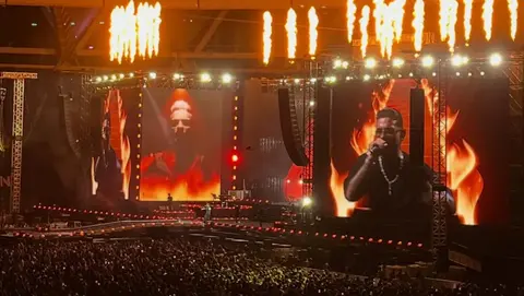 A performer sings on stage with large screens displaying visuals and flames above, while a crowd watches in an outdoor stadium.