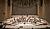 Empty concert hall stage with wooden seating, featuring a circular arrangement of music stands and chairs, surrounded by vertical wooden panels.