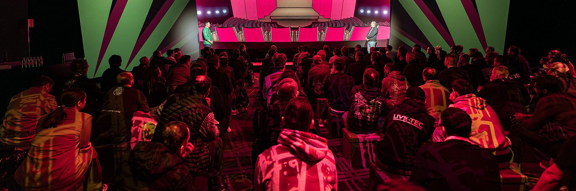 Audience seated in a dark venue, facing a brightly lit stage with colorful geometric screens and a panel of speakers.