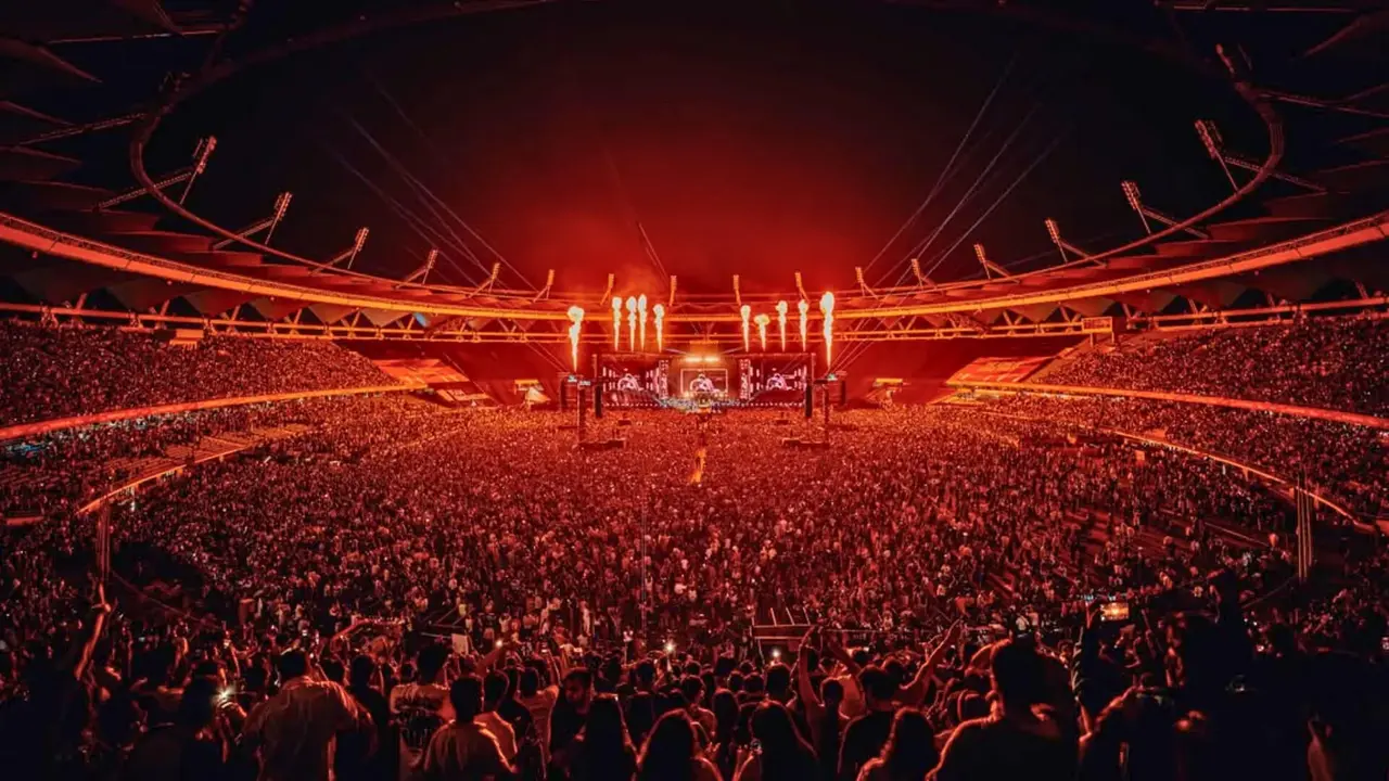 A large crowd gathers in a stadium during a concert, with bright lights and a vibrant atmosphere.