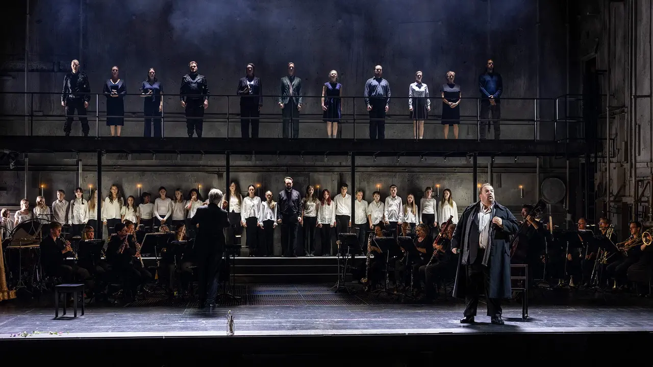 A theatrical performance featuring a conductor, an orchestra, and a choir on a multi-level stage, with performers in formal attire.