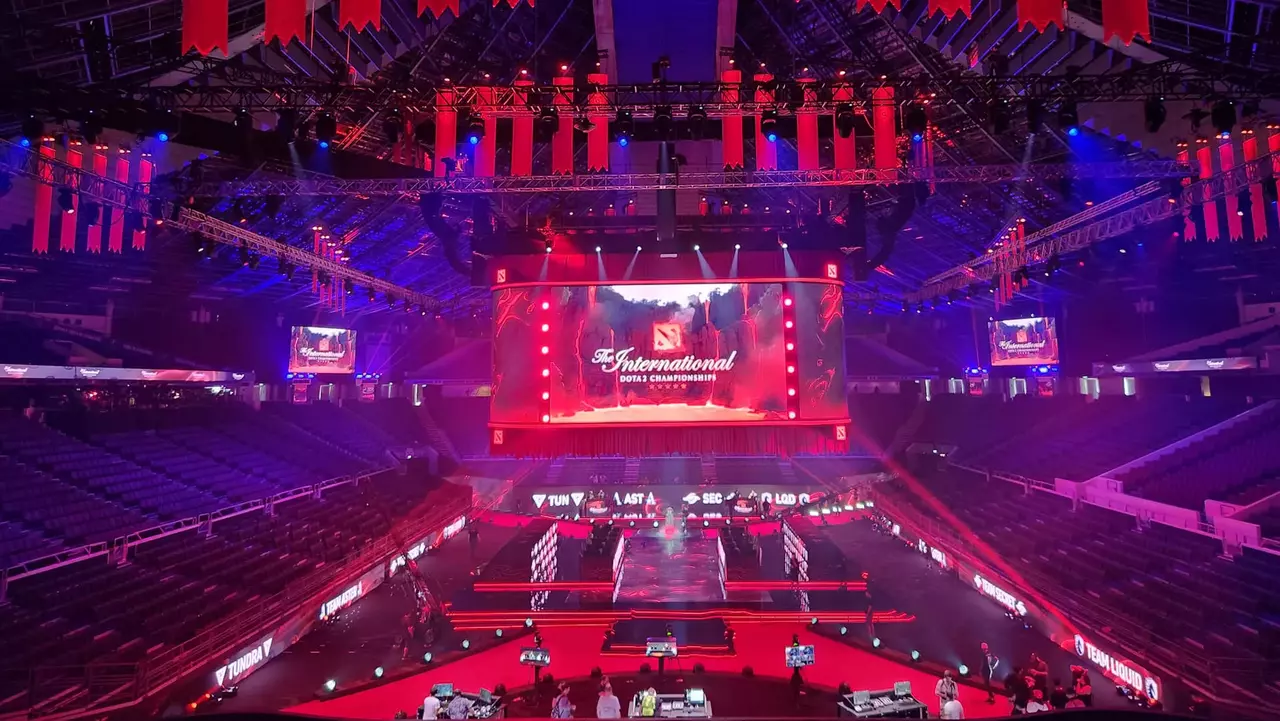 A large arena set up for an esports event, featuring a central stage with a screen displaying "The International" and vibrant lighting.
