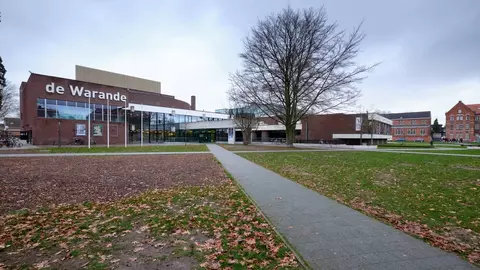 Modern building named "de Warande" with a glass facade, surrounded by a park area with fallen leaves and a pathway.
