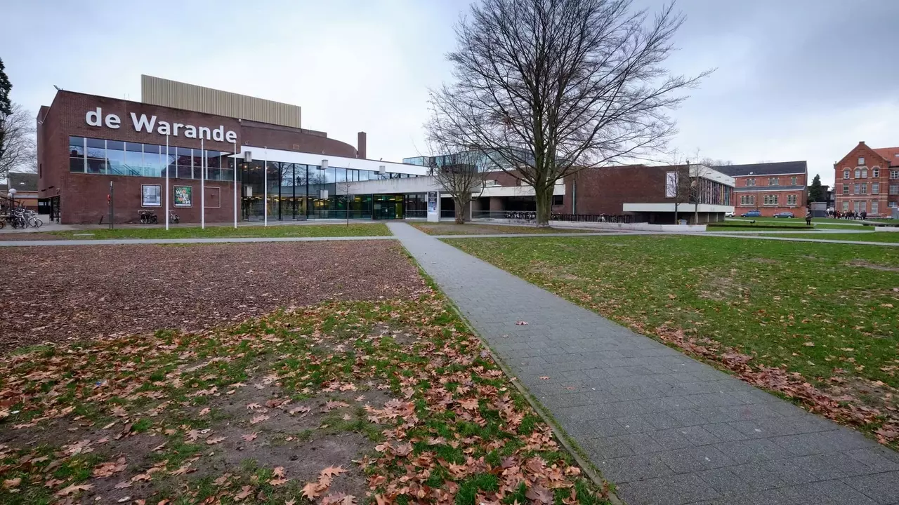 Modern building named "de Warande" with a glass facade, surrounded by a park area with fallen leaves and a pathway.