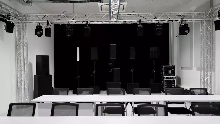A spacious room set up for an event, featuring rows of white tables and black chairs, with sound equipment and speakers arranged at the front.