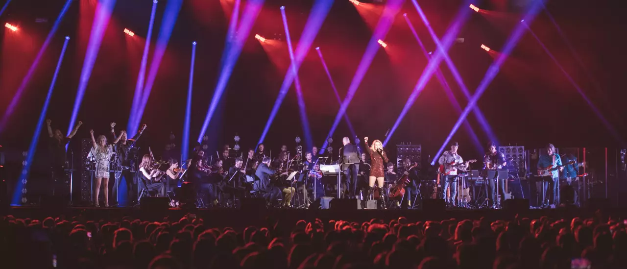 A live orchestra performance with musicians on stage, illuminated by colorful lights, while an audience watches from below.