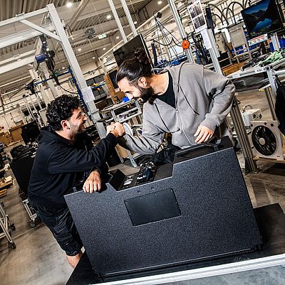 Two men are engaged in a friendly fist bump over a large black object in a factory setting, surrounded by equipment and workstations.