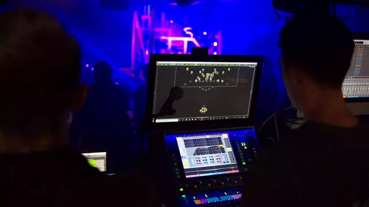 Two sound technicians operate digital mixing consoles in a dimly lit venue with colorful stage lighting in the background.