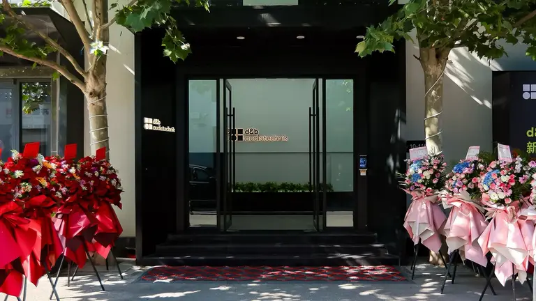 Entrance of a building adorned with floral arrangements on either side, featuring red and pink flowers, and a modern glass door.