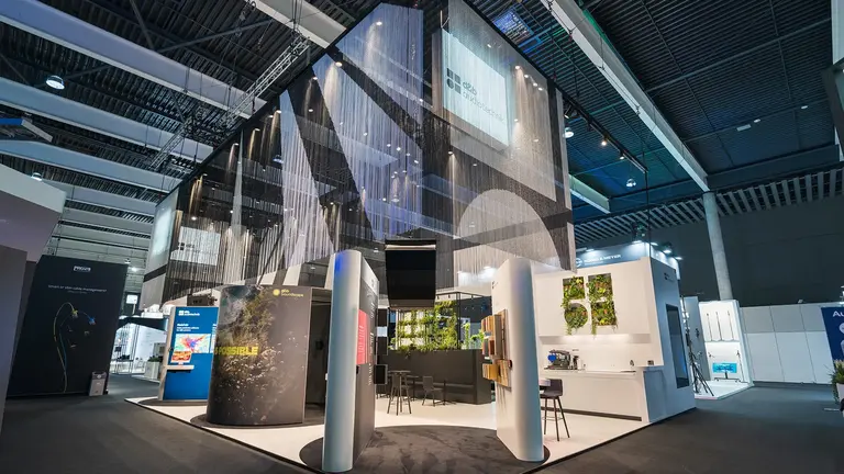 Moderner Messestand mit geometrischem Design, hängenden Elementen, Ausstellungsflächen und Sitzbereichen, in einem geräumigen Kongresszentrum eingerichtet.