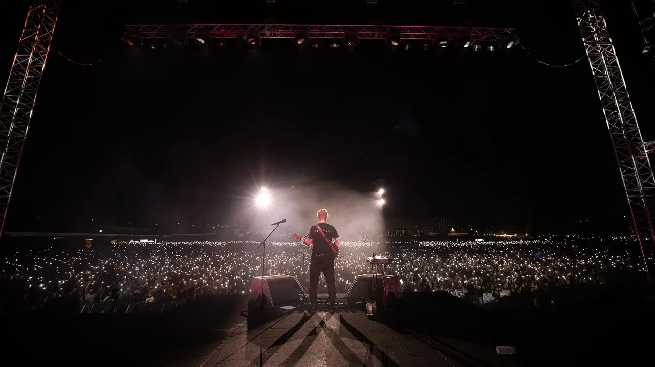A musician performs on stage, facing a large crowd illuminated by phone lights at night.
