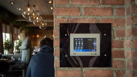 Control panel mounted on a brick wall in a restaurant, displaying settings with sliders and a digital clock. A person sits at a table nearby.