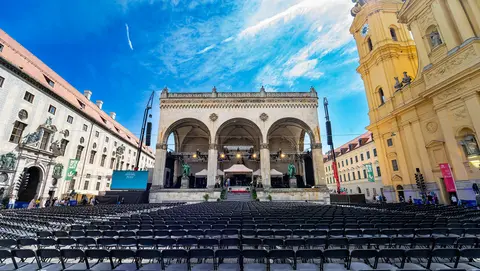 A large outdoor venue with rows of empty chairs set up in front of an ornate stage, flanked by historic buildings under a clear blue sky.