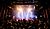 A crowd enjoys a live music performance on stage, illuminated by bright lights in a concert venue.