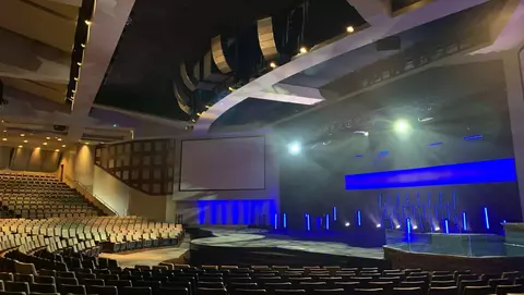 Interior of a modern auditorium featuring rows of seats, a stage with blue lighting, and large screens on the walls.