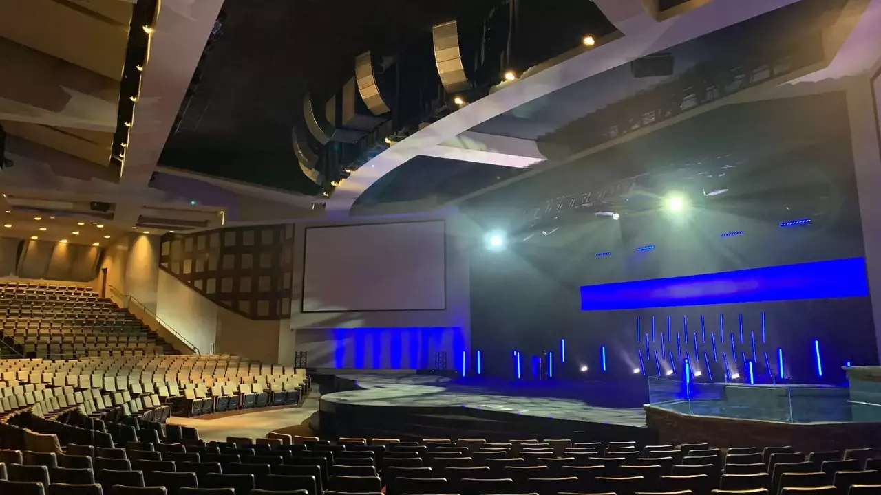 Interior of a modern auditorium featuring rows of seats, a stage with blue lighting, and large screens on the walls.