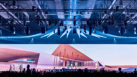 A large stage with a modern architectural backdrop is illuminated by bright lights, while a crowd watches a performance or presentation.
