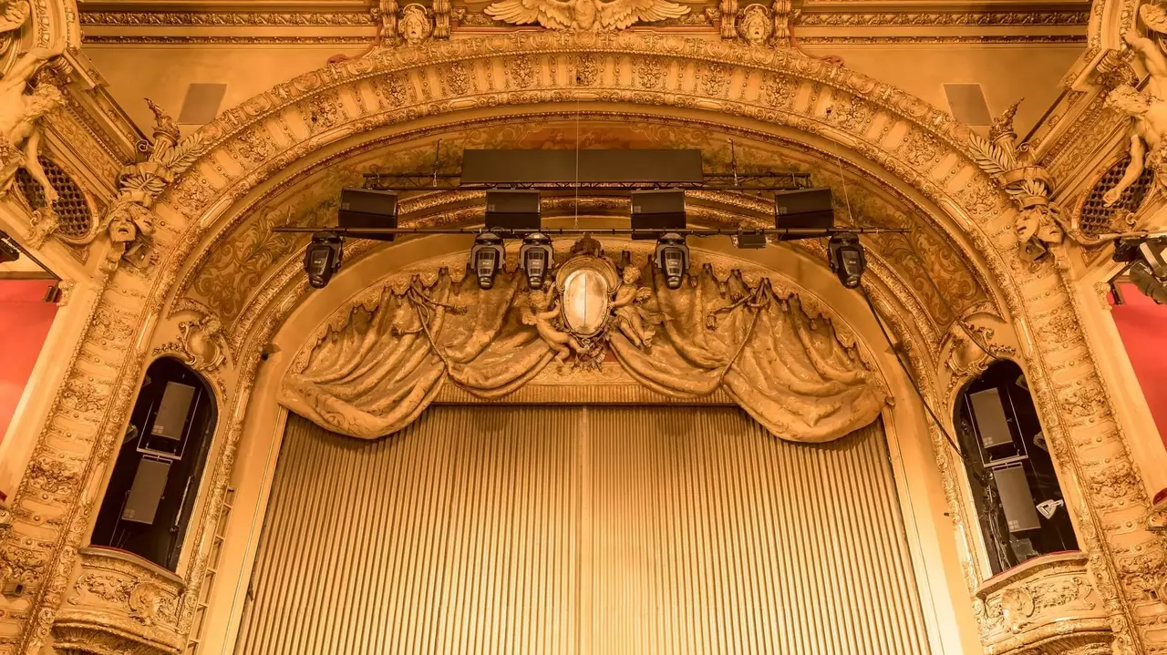 Ornate theater proscenium arch with decorative elements and curtains, featuring lighting fixtures above a striped backdrop.