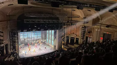 A theater performance with dancers on stage, illuminated by stage lights, while an audience watches from the seating area.