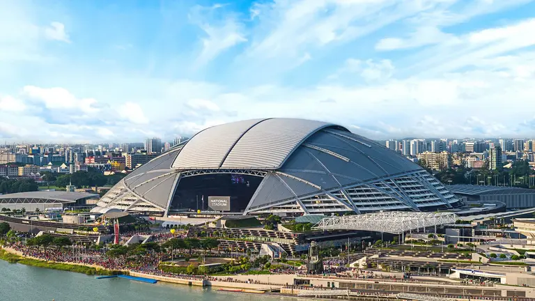 The National Stadium in Singapore features a distinctive domed roof and is surrounded by a waterfront area, with a crowd gathered outside.
