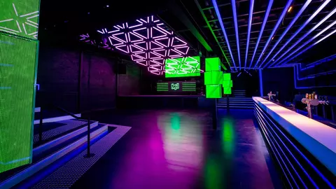 A modern nightclub interior featuring vibrant purple and green lighting, geometric ceiling designs, and a sleek bar area.