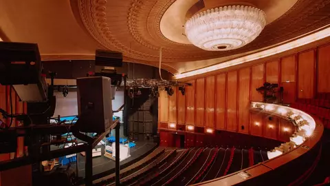 Interior view of a theater featuring a chandelier, wooden paneling, and seating arranged in a semi-circular layout.