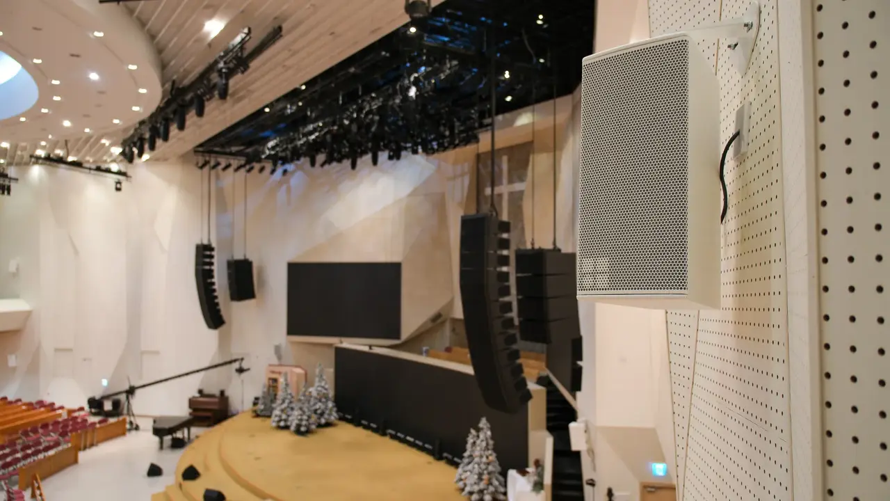 Auditorium interior featuring a stage with sound equipment, large screens, and Christmas trees arranged on the floor.