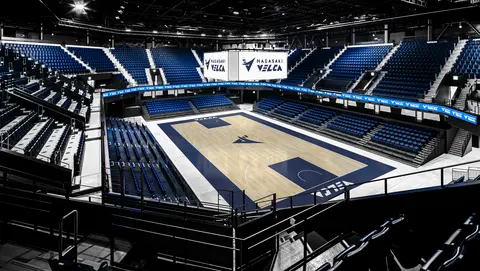 Indoor basketball arena featuring a polished court, surrounded by tiered seating and digital displays.