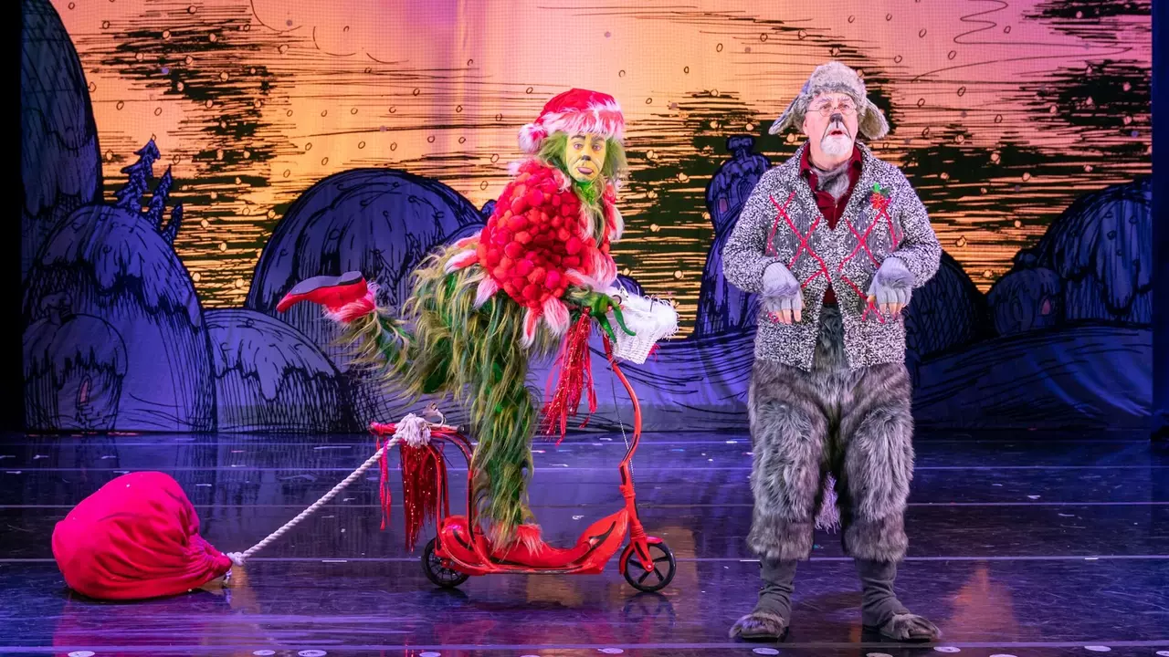 A character dressed as the Grinch rides a scooter while pulling a red sack, accompanied by another character in a festive outfit.