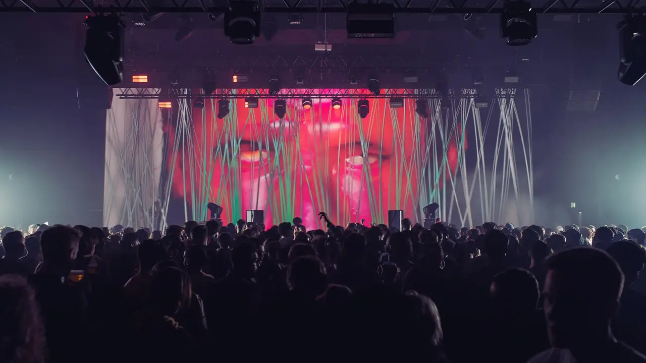 Crowd at a concert with vibrant lighting and a large screen displaying abstract visuals in the background.