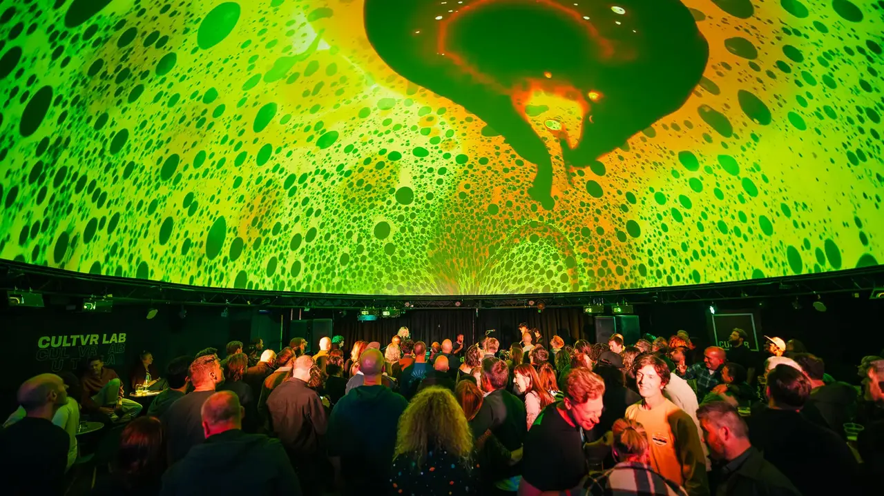 A crowd gathers under a vibrant, colorful projection on the ceiling, featuring abstract patterns in green and orange hues.
