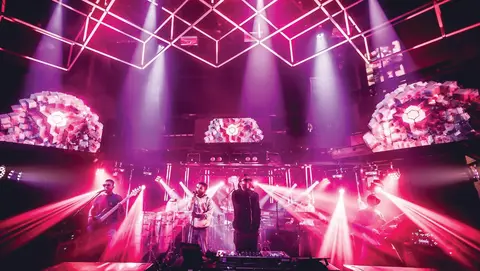 A band performs on stage with vibrant lighting and visual displays, featuring musicians playing instruments and singing.