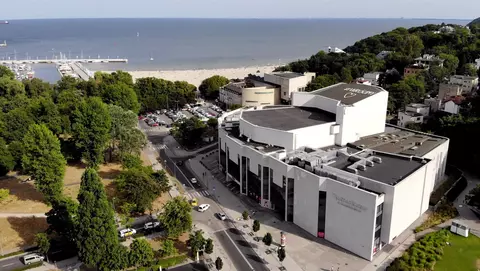 Aerial view of a modern building near the coast, surrounded by trees and a parking area, with a beach and ocean in the background.