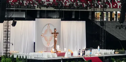 A stage setup featuring a large crucifix, white drapes, and seating arrangements, prepared for a religious event in a stadium.