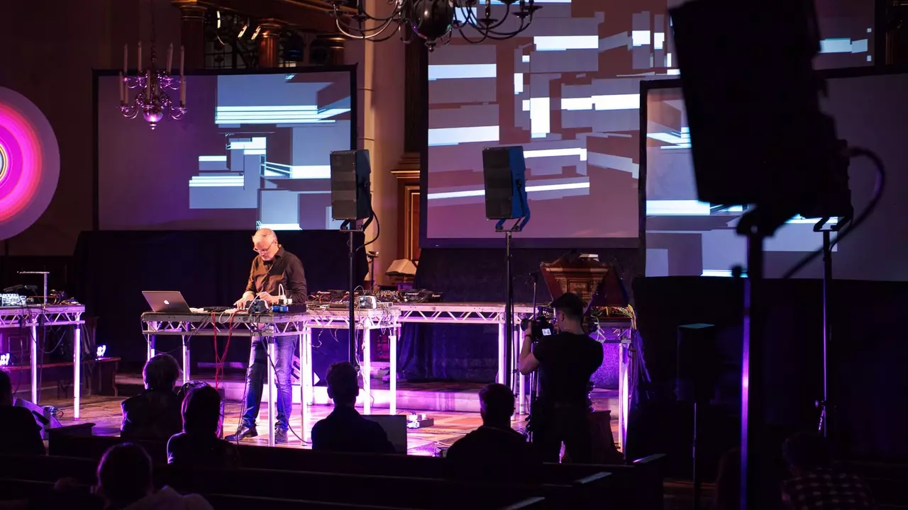 A performer stands at a table with electronic equipment, surrounded by screens displaying abstract visuals, while an audience watches.