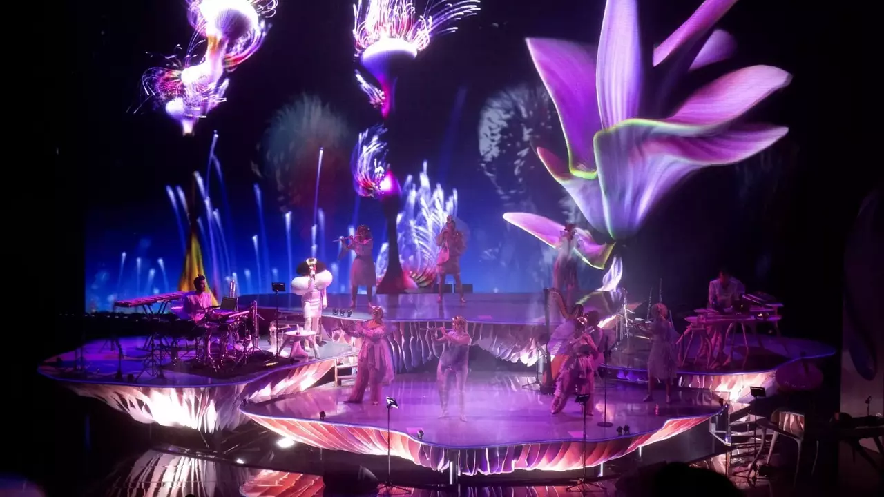 A vibrant stage performance featuring dancers and musicians surrounded by large, colorful floral projections and dynamic visual effects.